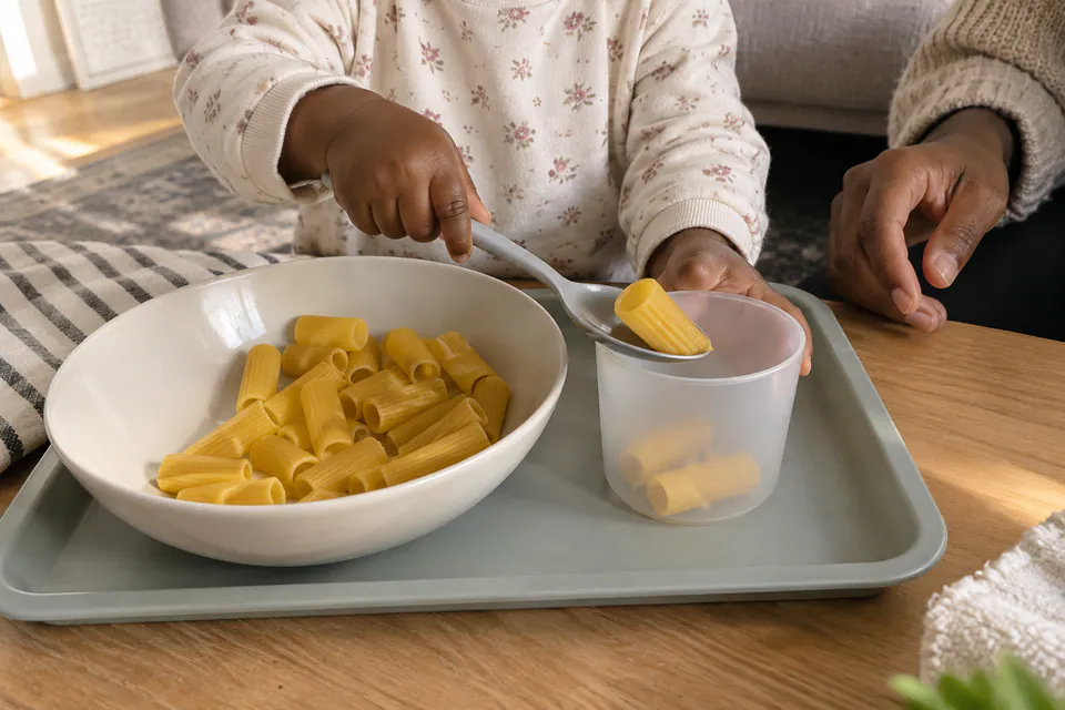 A child spooning dry pasta from a bowl into a cup.