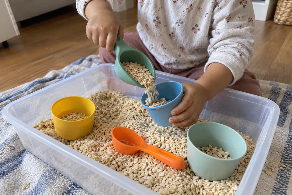 A child scooping dry oats into cups in a shallow sensory bin.