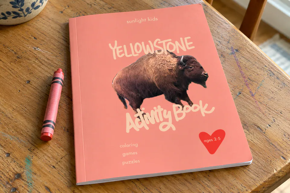 The pink Yellowstone Activity Book for ages 2–5 on a wooden table beside crayons.