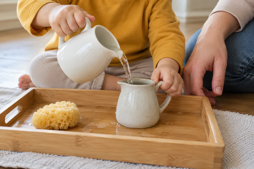 A child pouring water between two small pitchers on a tray beside a sponge.